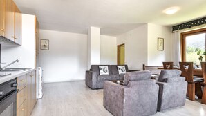 Eine moderne Küche mit Holzausstattung und einem eingebauten Backofen. Ein graues Sofasitzgefüge mit Kissen und ein Esstisch mit Stühlen sind vorhanden. Das Zimmer verfügt über ein großes Fenster mit Blick auf Grünflächen.