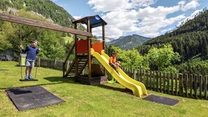 Ein Spielplatz mit Schaukel und Rutsche, umgeben von einem Holzzaun und üppigem Grün.