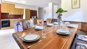 Ein moderner Essbereich mit einem Holztisch, gedeckt für sechs Personen, umgeben von Stühlen. Im Hintergrund ist eine Küche mit Holzschränken und einer Mikrowelle erkennbar.