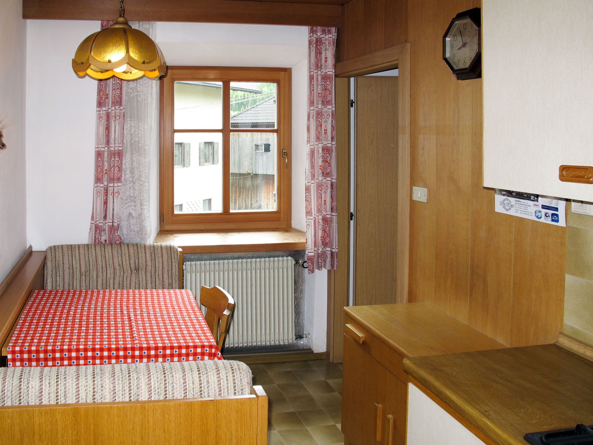 Una stanza accogliente con un letto di legno, un tavolo con tovaglia a scacchi rossi, una finestra con tende a motivi, un termosifone e un mobiletto in legno.