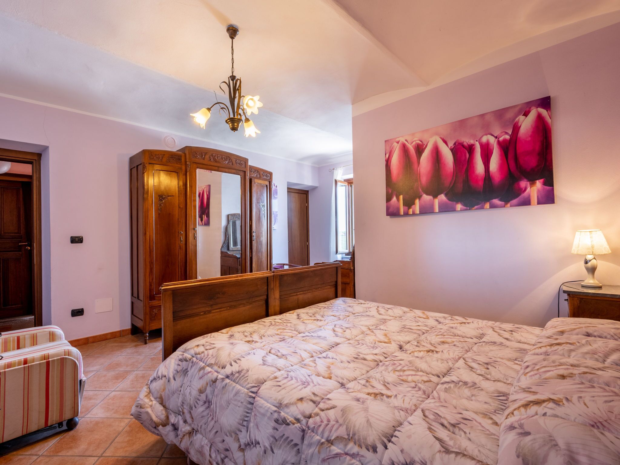 Une chambre à coucher avec un grand lit, une armoire en bois, un miroir et un tableau représentant des fleurs accroché au mur.