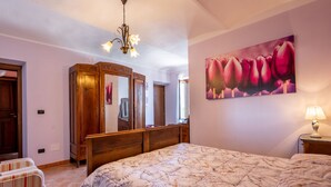 Une chambre à coucher avec un grand lit, une armoire en bois, un miroir et un tableau représentant des fleurs accroché au mur.