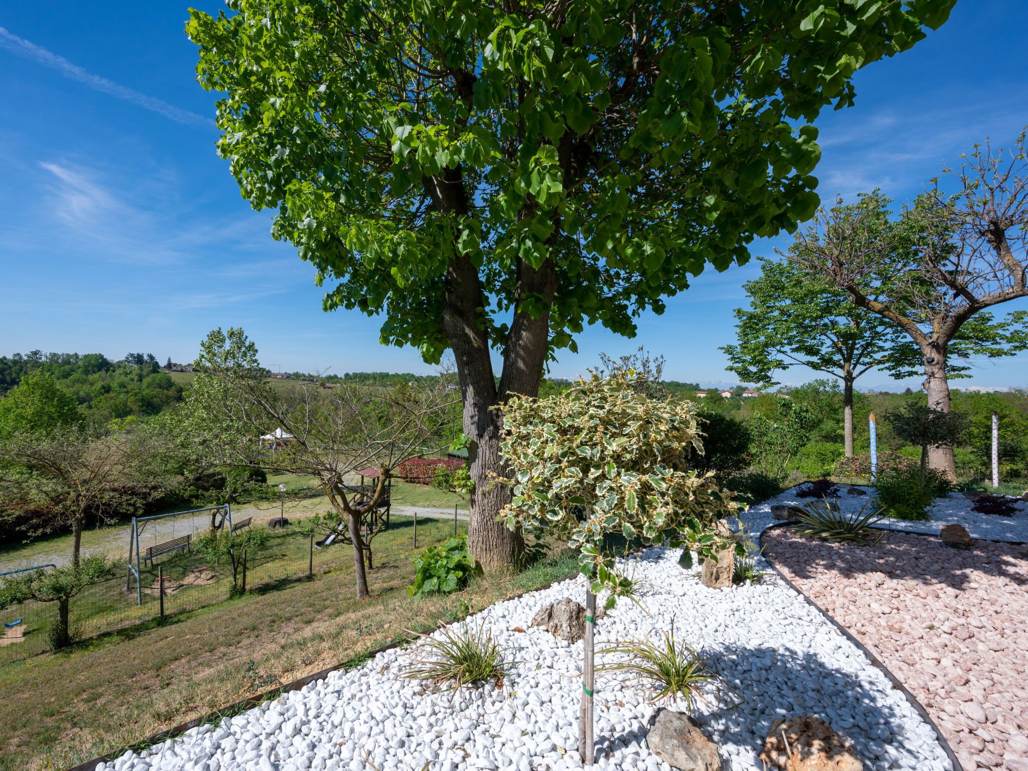 Un jardin aménagé avec un grand arbre, un chemin recouvert de gravier blanc et une vue sur un village situé au loin.