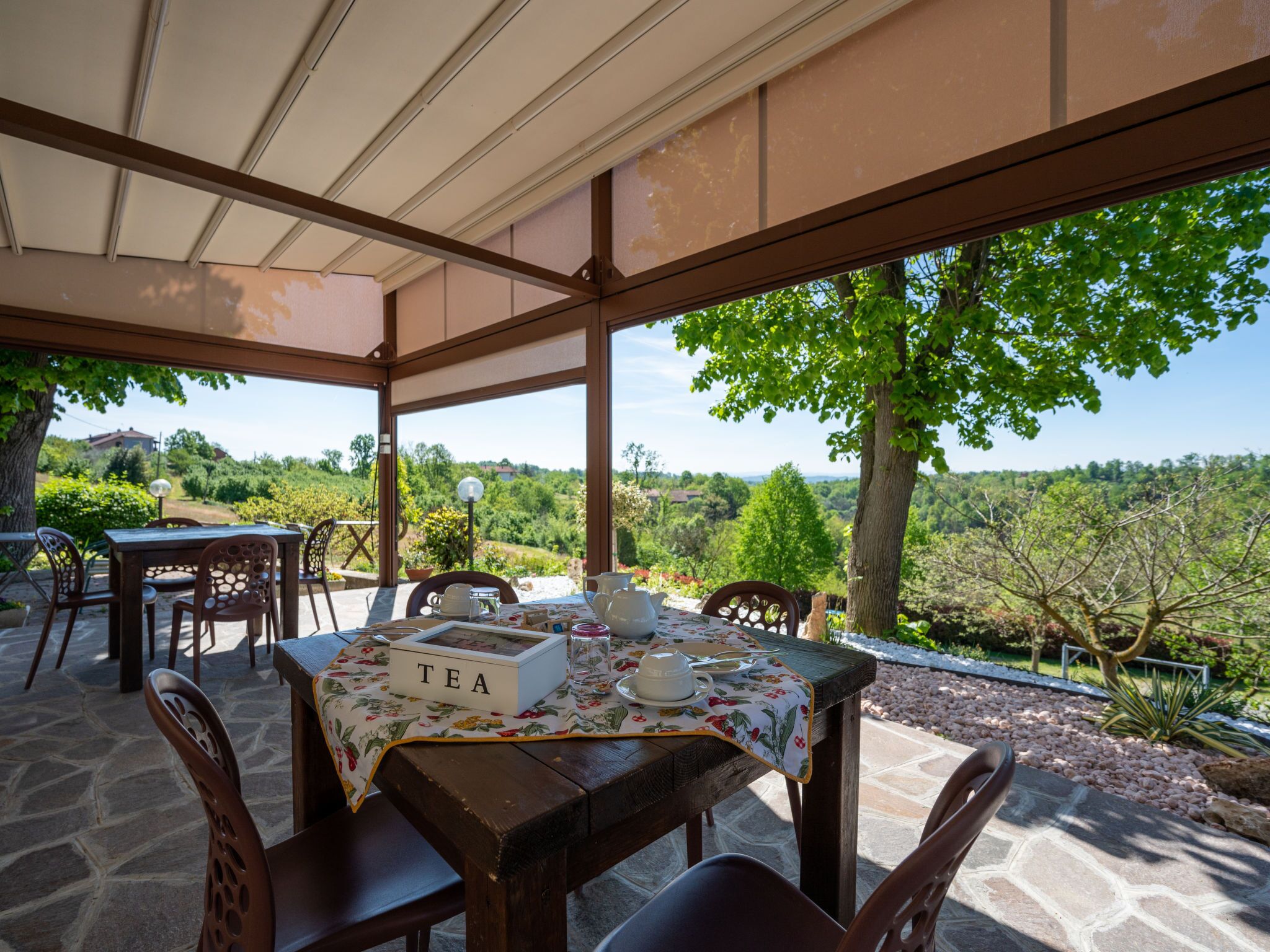 An outdoor dining area with a table set for tea, surrounded by stone flooring and wooden chairs.