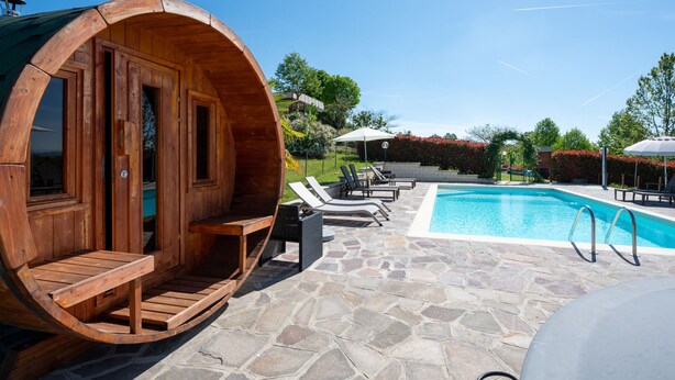 Un sauna en forme de tonneau, fait de bois, situé à côté d’une piscine dont le sol est recouvert de pierres, avec des chaises longues et un ciel d’un bleu limpide.