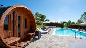 Un sauna en forme de tonneau, fait de bois, situé à côté d’une piscine dont le sol est recouvert de pierres, avec des chaises longues et un ciel d’un bleu limpide.