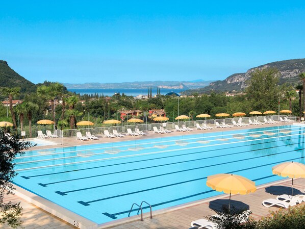 A swimming pool with multiple lanes, sun loungers, and umbrellas, surrounded by greenery and a scenic view of hills and the sea.