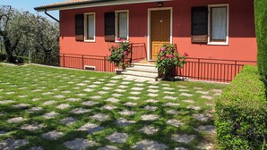 A red building with a tiled roof, a front door, and two windows with shutters. There is a small garden with a lawn and a paved area with stepping stones. - Apartment Gianni by Interhome (Torri del Benaco)