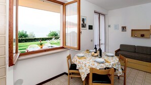 A dining area with a table set for four, a window with an open shutter, a sofa, and a small shelf with items. - Apartment Gianni by Interhome (Torri del Benaco)