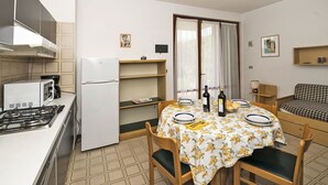 Une cuisine compacte équipée d’une cuisinière à gaz, d’un four à micro-ondes et d’un réfrigérateur. Une table à manger avec une nappe à motifs floraux et des chaises est placée au centre de la pièce.