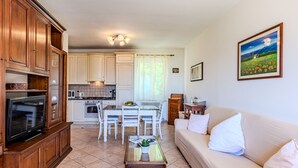 A living room with a sofa, a dining table with chairs, a kitchen area with cabinets and appliances, and a painting on the wall.