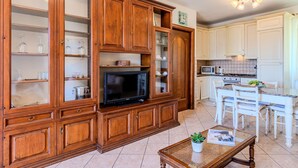 A spacious living area with wooden cabinetry, a flat-screen TV, and a dining area with white chairs and a kitchen in the background.