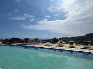 Un espace piscine avec des chaises longues et des tables, un ciel dégagé et une vue lointaine sur la mer et les collines.