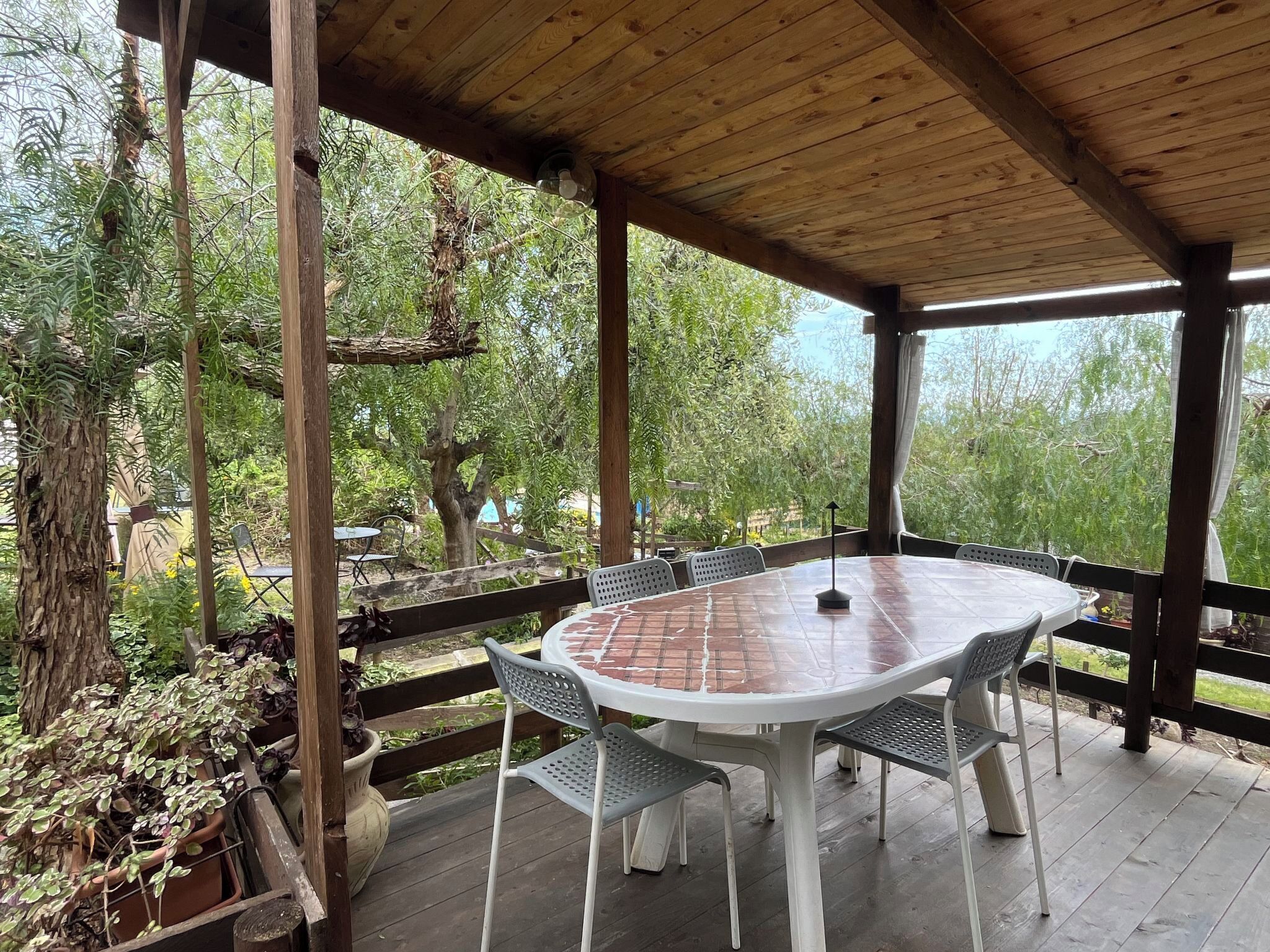 A covered patio with a round table and four chairs, surrounded by wooden beams and potted plants.