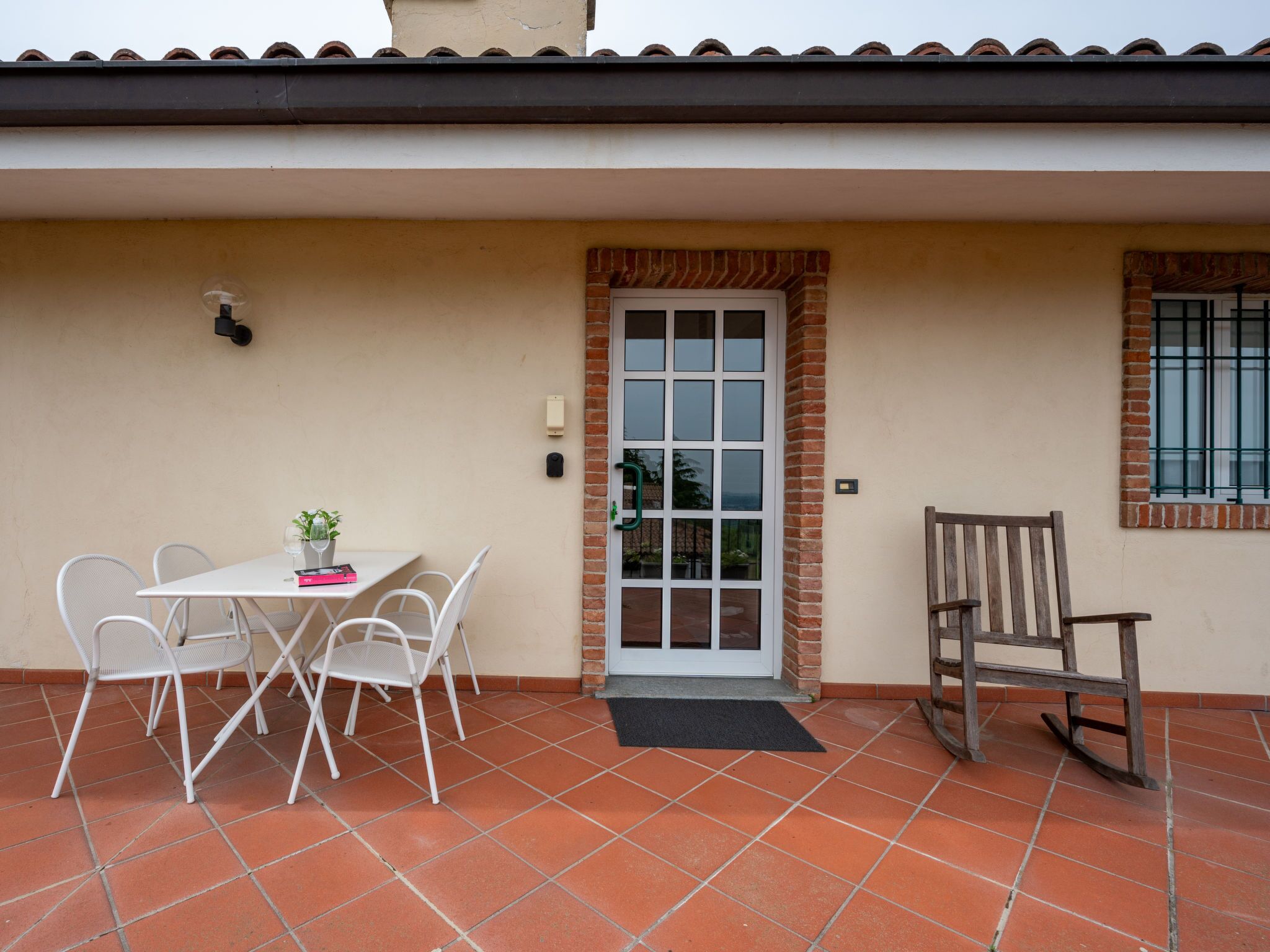 Un patio con una mesa y sillas blancas, una mecedora de madera, y una puerta con un panel de vidrio.