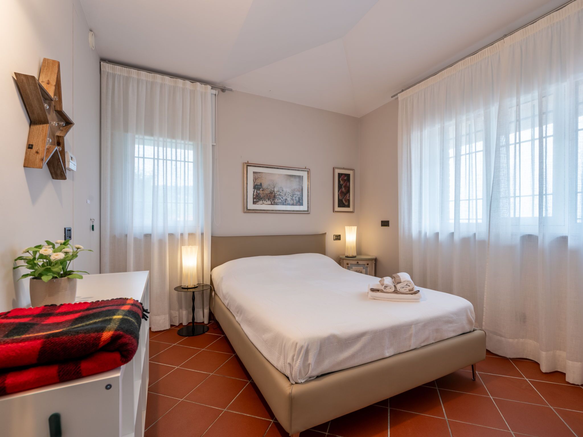 Une chambre d’hôtel avec un lit, une table de chevet, une lampe, une plante et une fenêtre avec des rideaux.