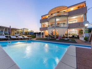 Un edificio de dos pisos con piscina, mobiliario exterior y una zona cubierta con plantas.