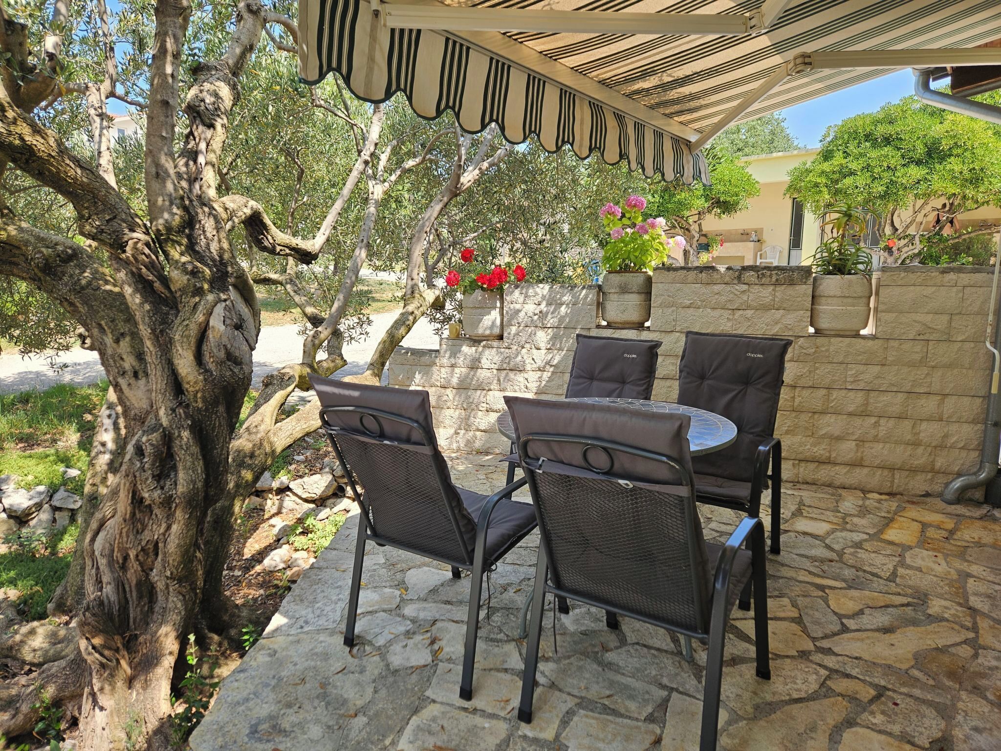An outdoor patio with stone flooring, a pergola with striped awning, and a table with four chairs.