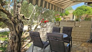 Une terrasse extérieure avec un sol en pierre, une pergola surmontée d’une marquise à rayures et une table avec quatre chaises.