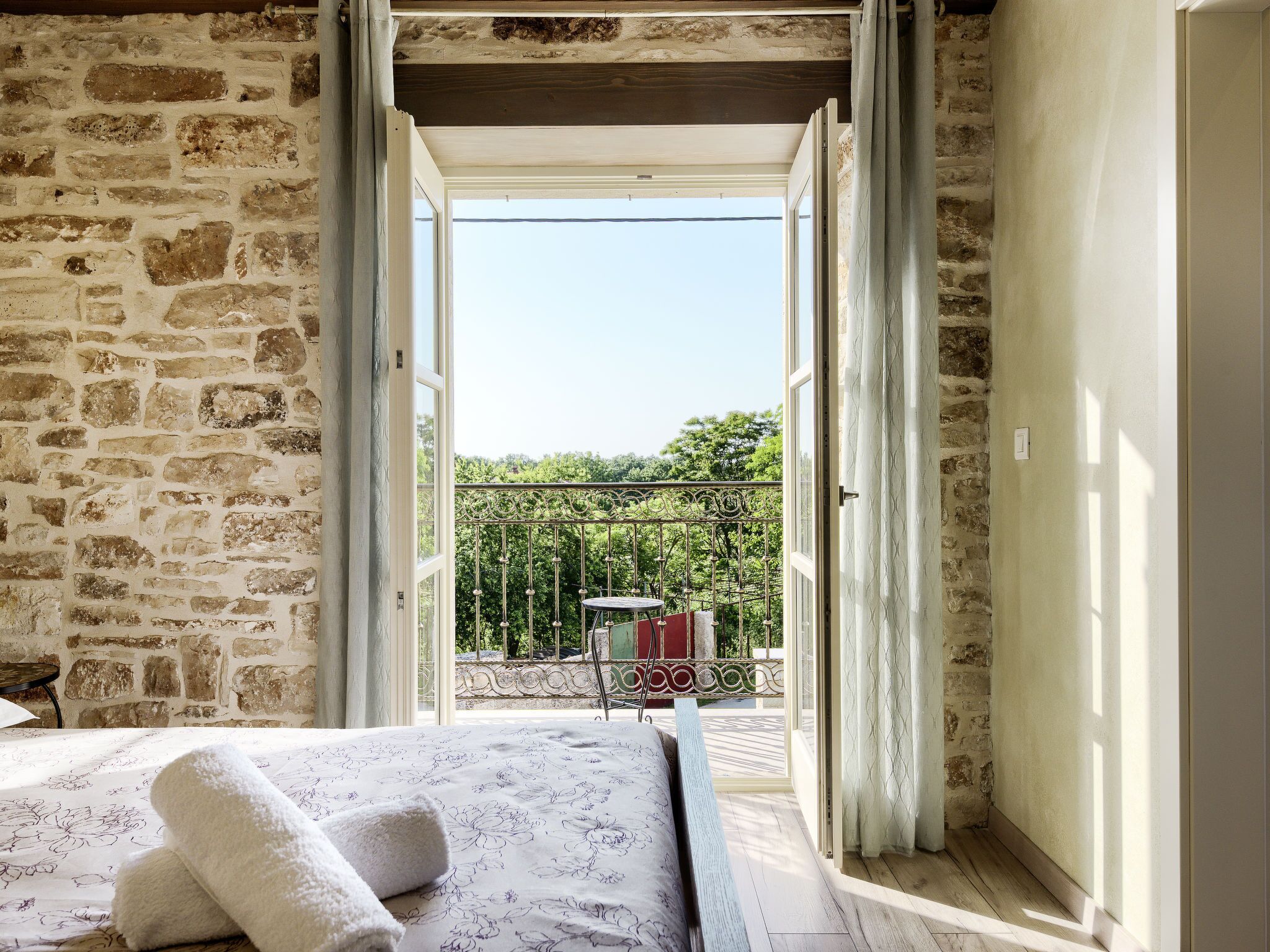 A room with a stone wall, a bed with a floral bedspread, and a view of a balcony with a table and chairs.
