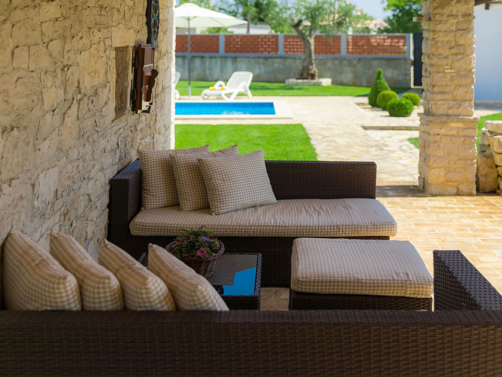 A wicker sofa with checkered cushions, a small table with a basket of flowers, and a view of a swimming pool and garden in the background.