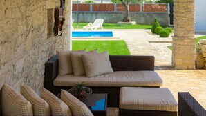 Un canapé en osier avec des coussins à carreaux, une petite table sur laquelle est posé un panier de fleurs, et en arrière-plan, une vue sur une piscine et un jardin.