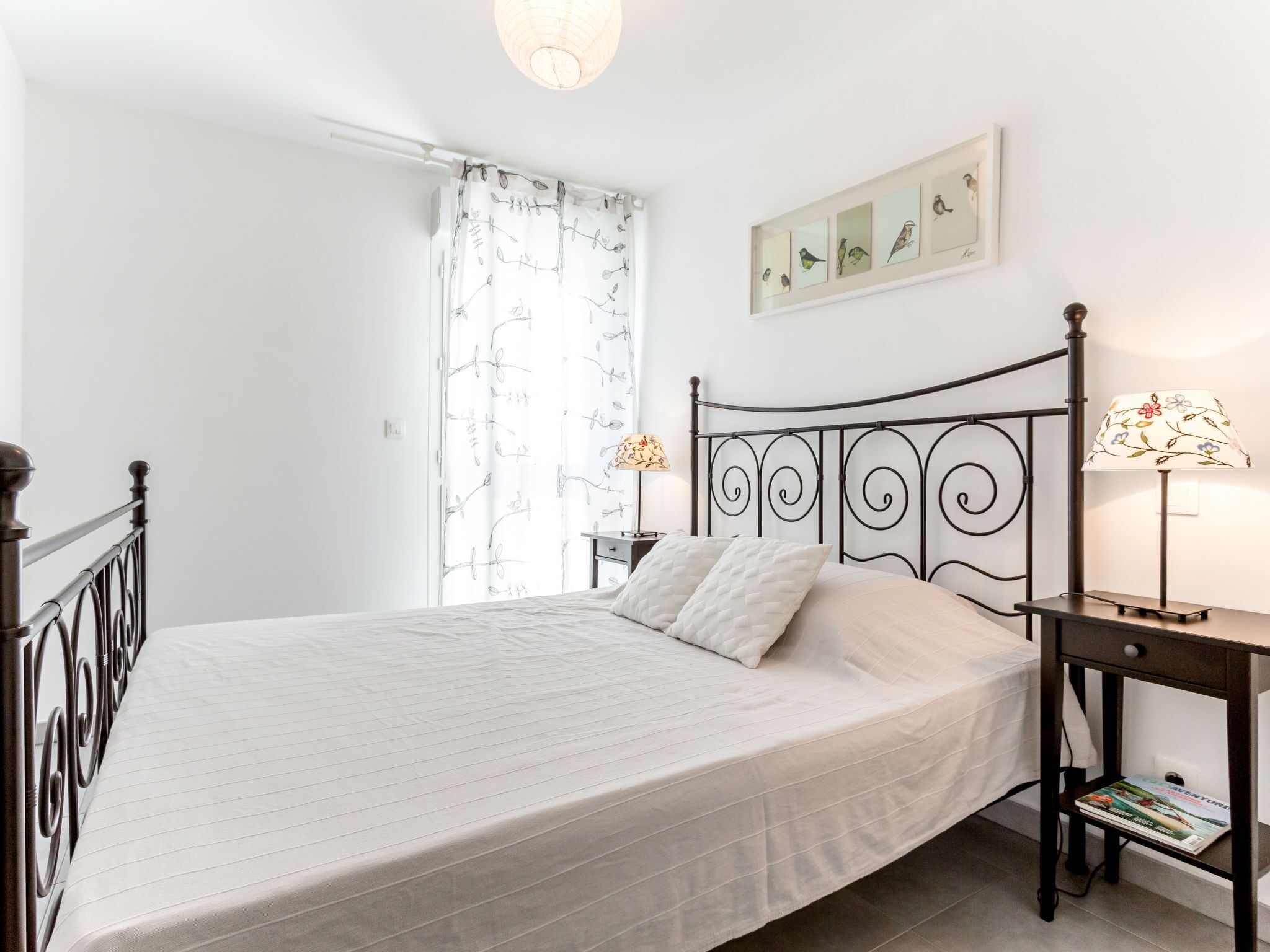 Un lit bien fait, recouvert d’une couvre-lit blanche et agrémenté d’oreillers, avec une tête de lit en métal noir, une table de chevet sur laquelle se trouve une lampe, et un tableau représentant des oiseaux accroché au mur.