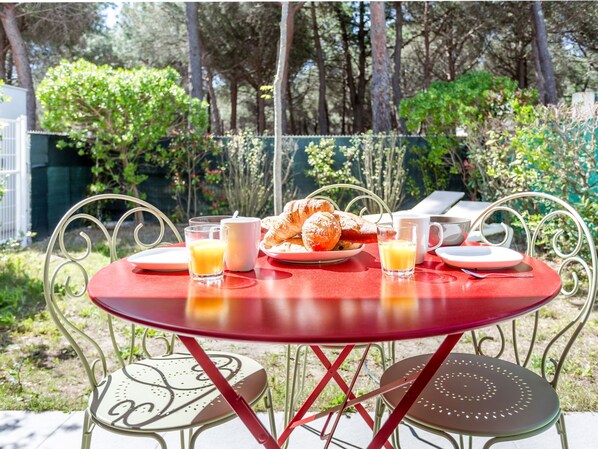 A red table with breakfast items including croissants, juice, and coffee, set outdoors with metal chairs and a garden background.