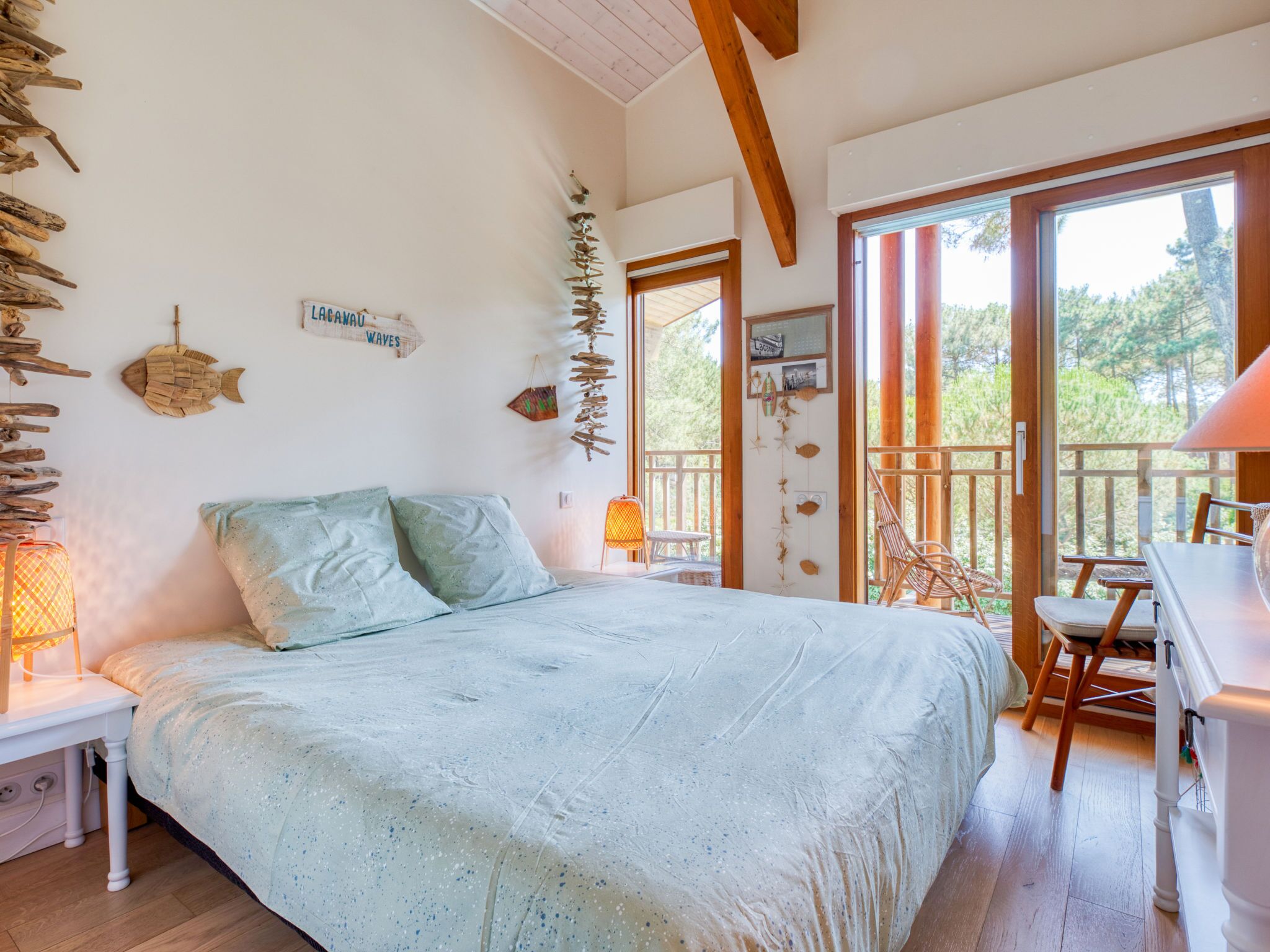 Un dormitorio con cama, mesita de noche, escritorio y silla. Hay vigas de madera en el techo y una ventana con vistas a árboles.