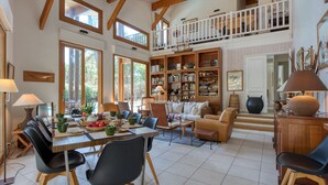 Un amplio salón con una mesa de comedor, sillas y un sofá. Hay estanterías llenas de libros y objetos decorativos. Un balcón da a este espacio.