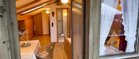 A wooden-framed room with a dining table and chairs, a glass door leading to a bathroom, and a window with curtains.