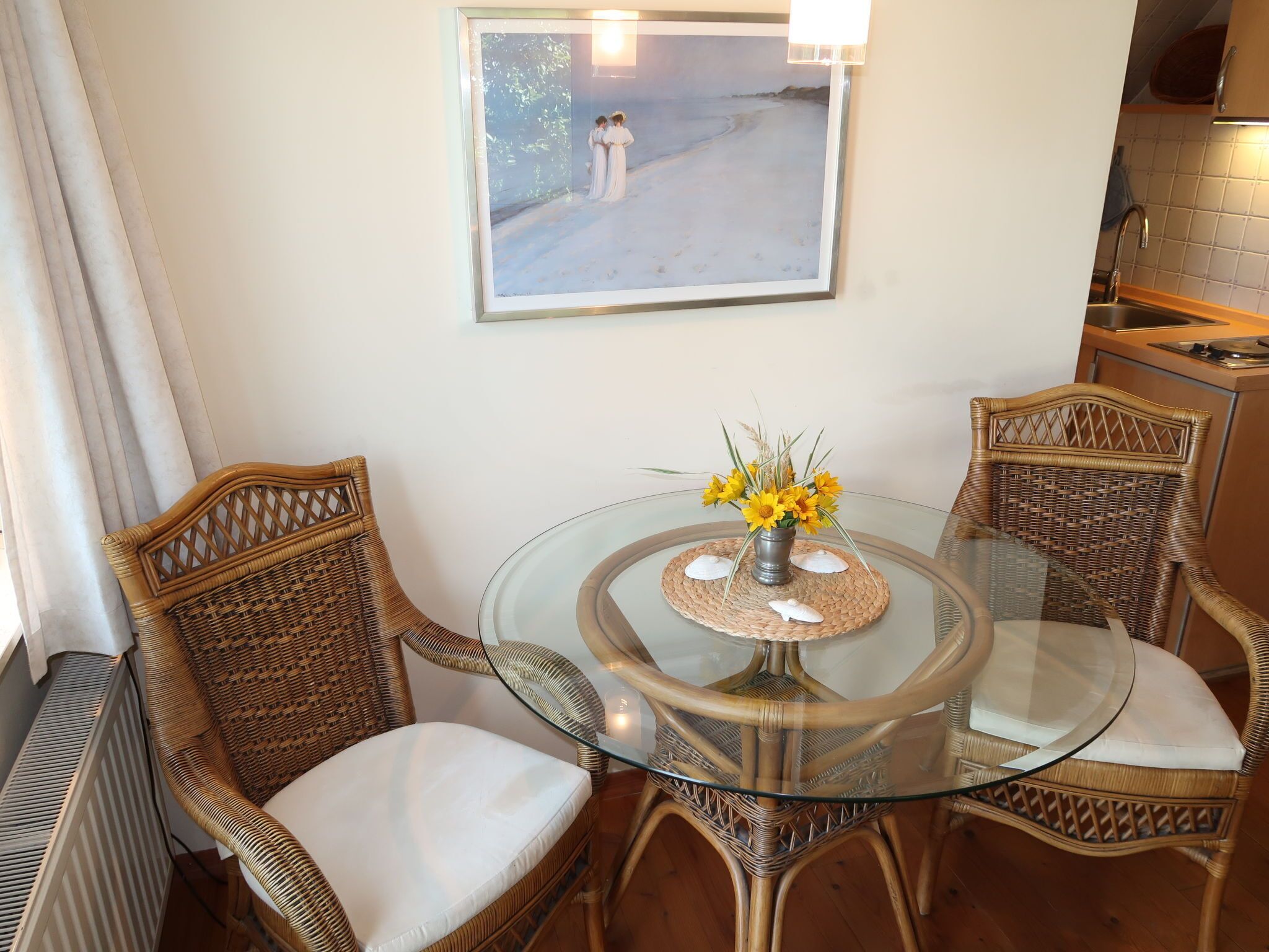 A dining area with wicker chairs, a glass table, and a framed painting on the wall.