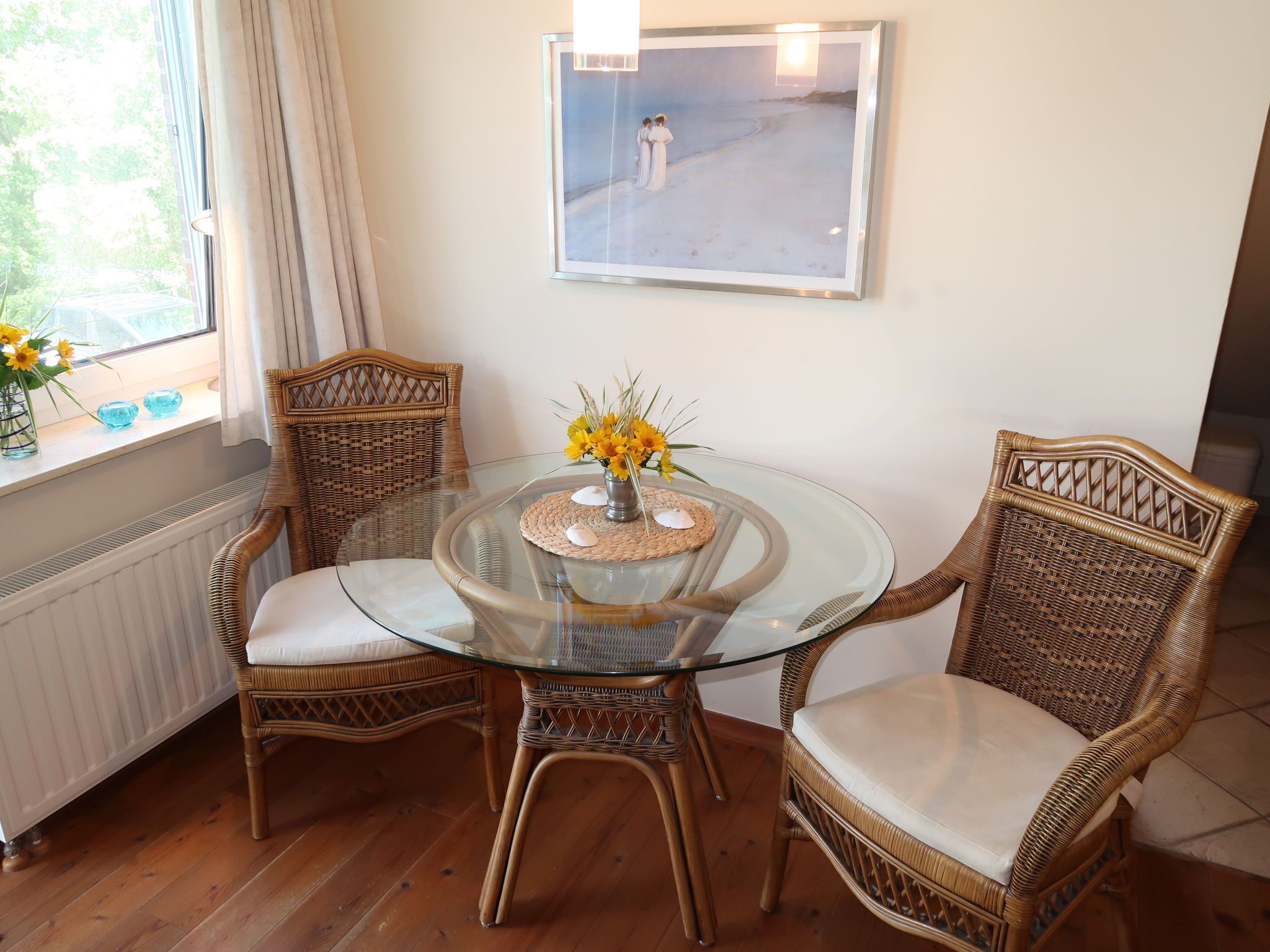 A dining area with a glass table surrounded by wicker chairs, a framed painting on the wall, and a window with curtains.