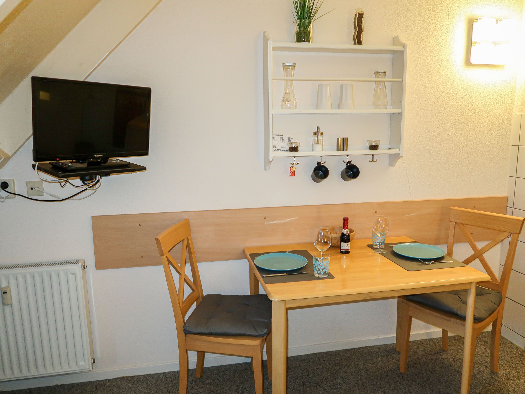 A small dining area with a wooden table and chairs, a television mounted on the wall, and a shelf with kitchen items.