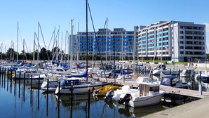 Un port esportiu amb diverses embarcacions amarrades, un edifici gran amb múltiples pisos i balcons, i un cel blau i clar.