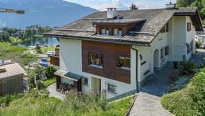 Una casa moderna con techo inclinado, sección de madera y vista a un lago y montañas al fondo.