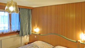 Une pièce avec un mur lambrissé en bois, un lit avec une literie à motifs floraux, un luminaire suspendu et une fenêtre avec des rideaux.