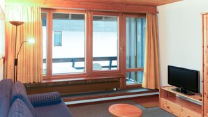 A room with a sofa, a small wooden table, a television on a wooden stand, and large windows with curtains.