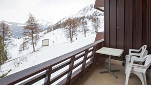 Un balcó amb vistes a muntanyes cobertes de neu, una barana de fusta i una taula amb dues cadires de plàstic blanc.