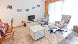 A modern living room with a flat-screen TV, two armchairs, a sofa, a coffee table, and a dining table with chairs.