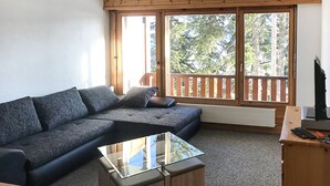 En moderne stue med en mørkegrå hjørnesofa, et sofabord med glasplade og et træskrivebord med en computer. Store vinduer giver udsigt til en balkon med gelænder af træ.