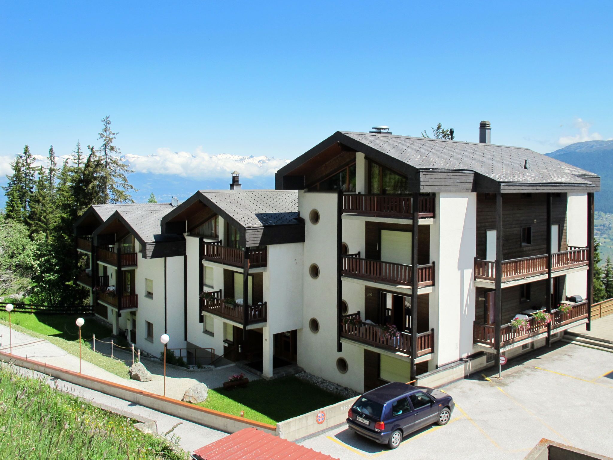 Un hotel moderno de varios pisos con balcones, un estacionamiento y vistas a montañas.