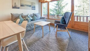 Een gezellige kamer met een houten plafond, een bank met kussens, een tafel, een stoel en grote ramen met uitzicht op bomen.