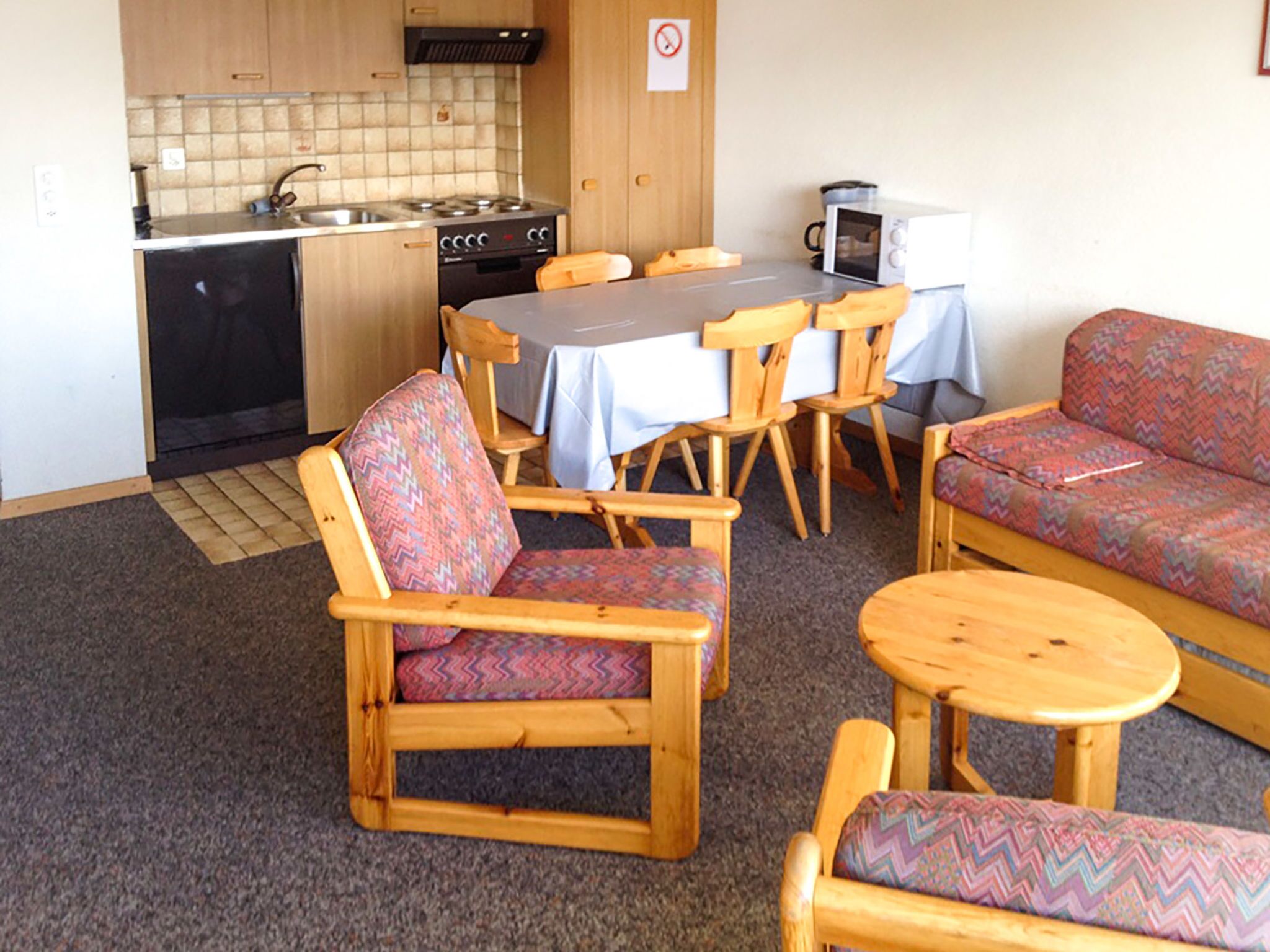 A compact kitchen with a stove, sink, and microwave. There are wooden chairs and a sofa with patterned cushions.
