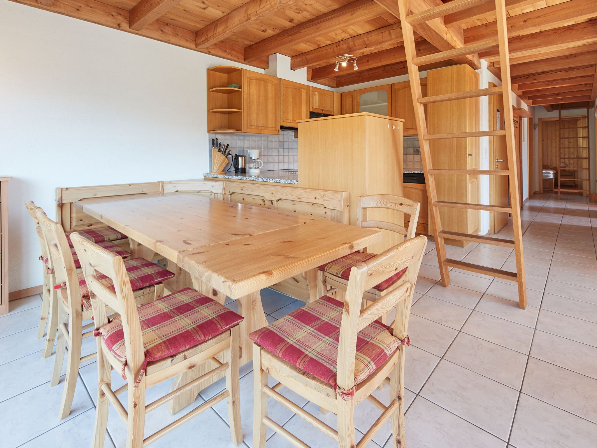Uma mesa de jantar de madeira com seis cadeiras, uma área de cozinha com armários de madeira e uma escada de madeira.