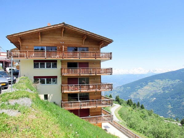 A multi-story wooden building with balconies, located on a hillside with a view of a mountainous landscape.