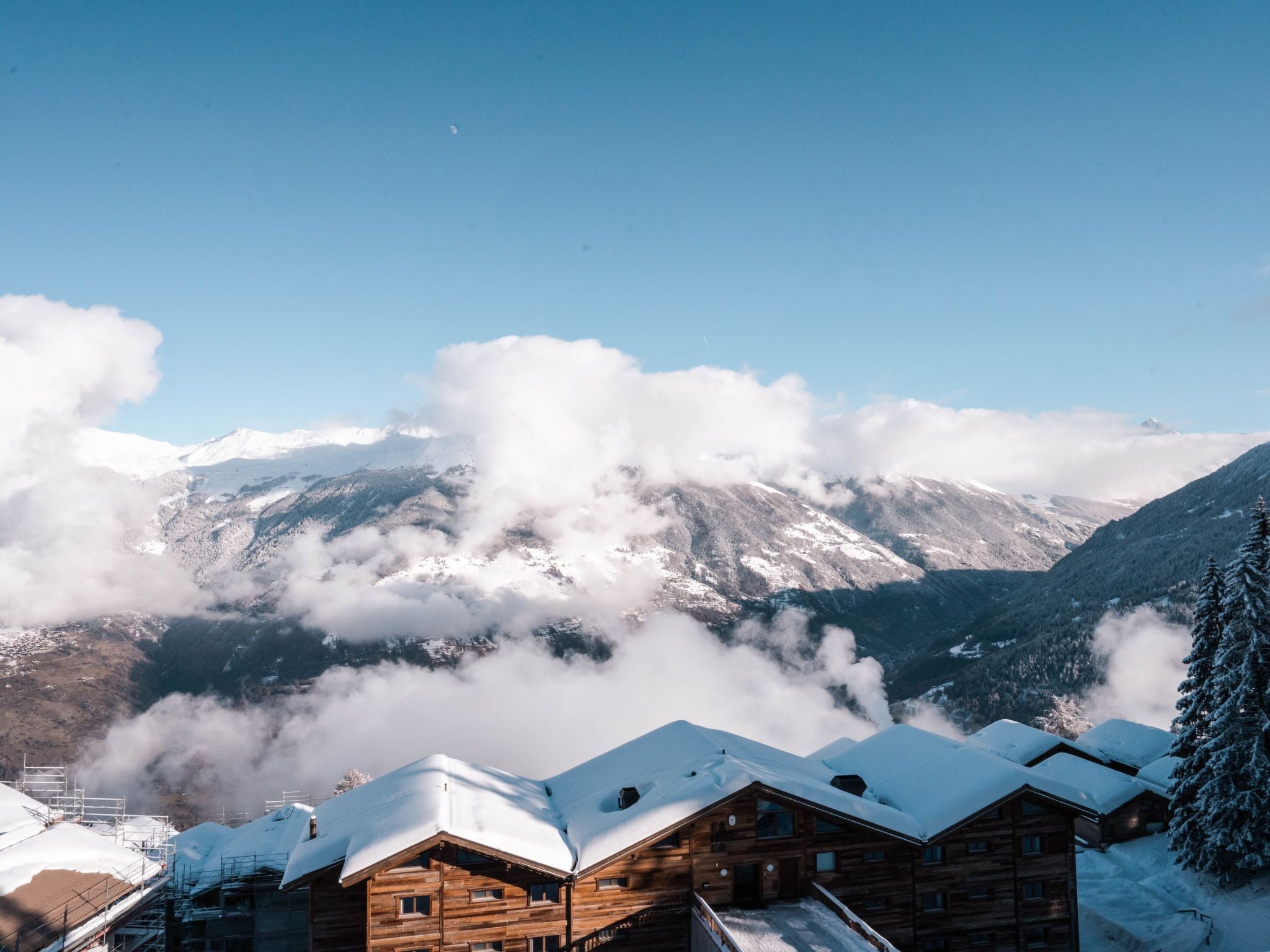 Una cabaña de madera con techo nevado, rodeada de montañas y nubes.