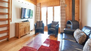 A cozy living room with a wooden ceiling, a fireplace, a sofa, two armchairs, a TV on a wooden cabinet, and a red rug.