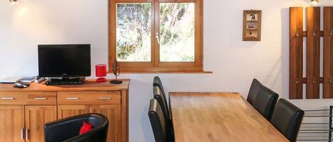 A dining area with a wooden table and chairs, a television on a cabinet, and a window with a view of trees.