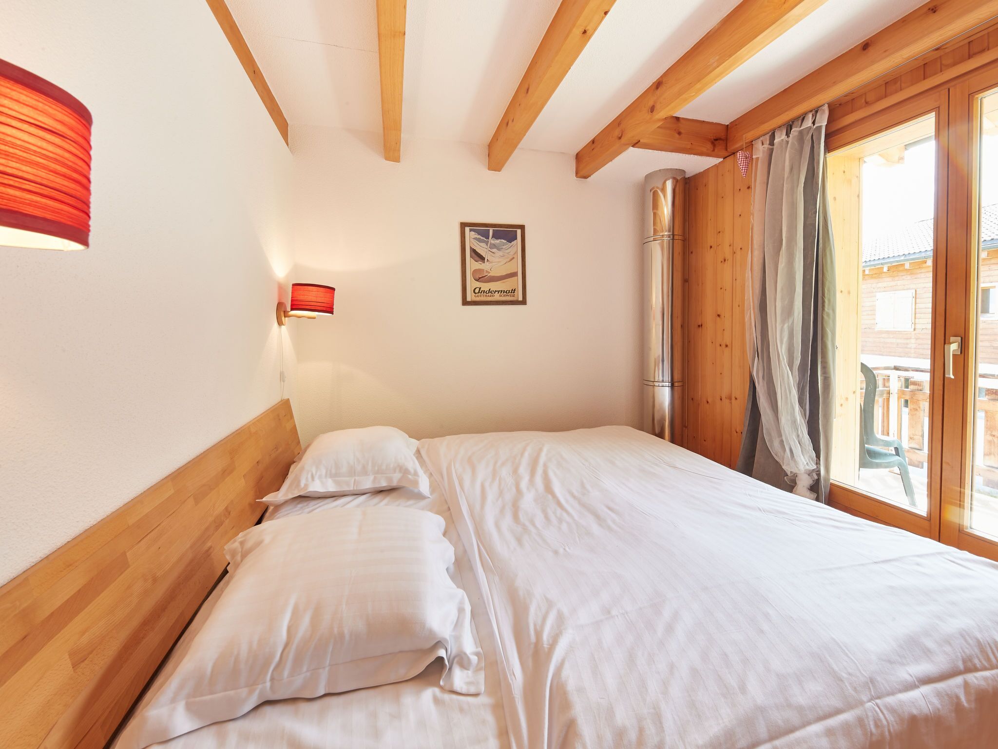 A bedroom with a wooden headboard, a bed with white linens, a wall-mounted lamp, a framed picture, and a window with curtains.